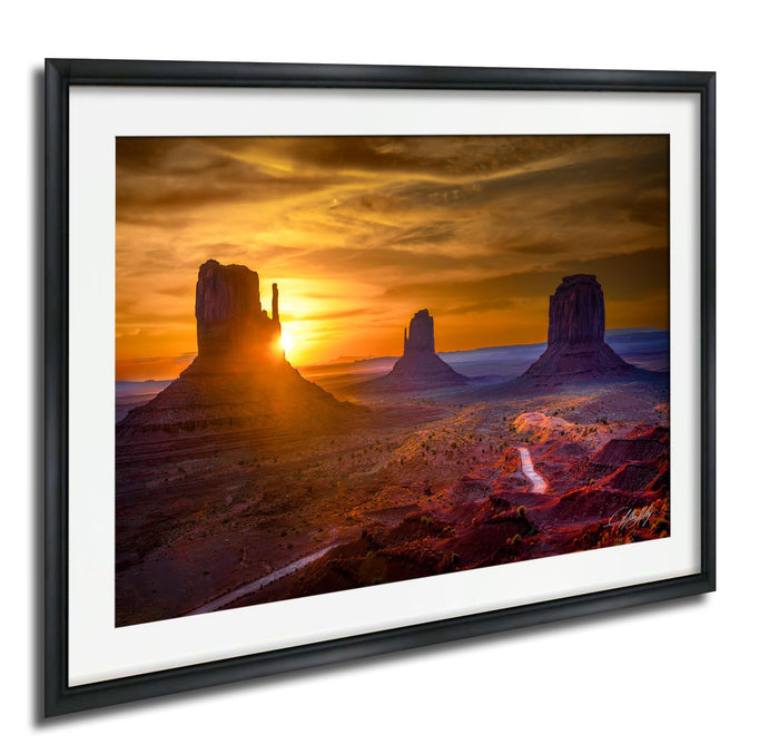 A framed photograph depicting the iconic rock formations of Monument Valley at sunset, with the sun setting behind the mountains and casting a warm, golden glow over the landscape.