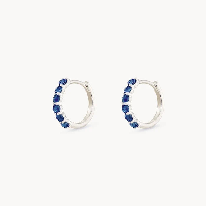 Side view of a pair of silver hoop earrings featuring a vertical channel setting of seven rose-cut deep london blue topaz gemstones, set against a plain off-white background.