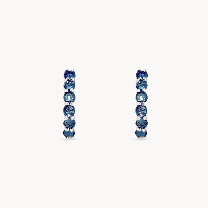 A pair of silver hoop earrings featuring a vertical channel setting of seven rose-cut deep london blue topaz gemstones, set against a plain off-white background.