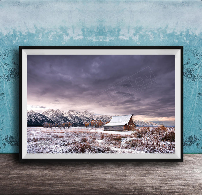 A framed photograph depicting a snowy mountain landscape with a wooden barn in the foreground, set against a cloudy sky.