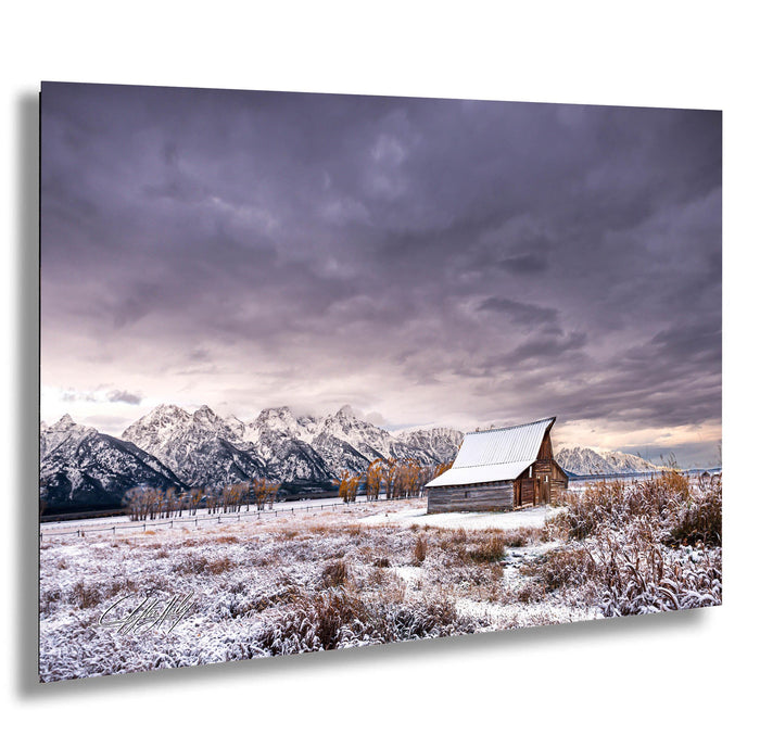 A snowy field with a wooden barn in the foreground, set against a backdrop of snow-capped mountains under a dramatic cloudy sky.
