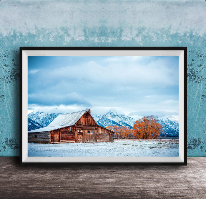 A large wooden barn with a sloping roof stands in a snowy field, with snow-capped mountains visible in the background.