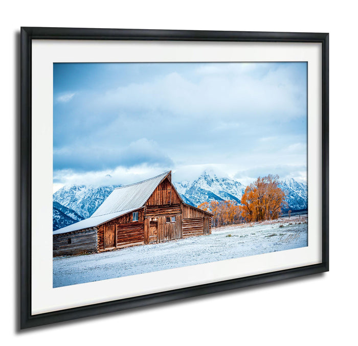 A rustic wooden barn with a sloping roof stands in a snowy landscape, with snow-capped mountains in the background.