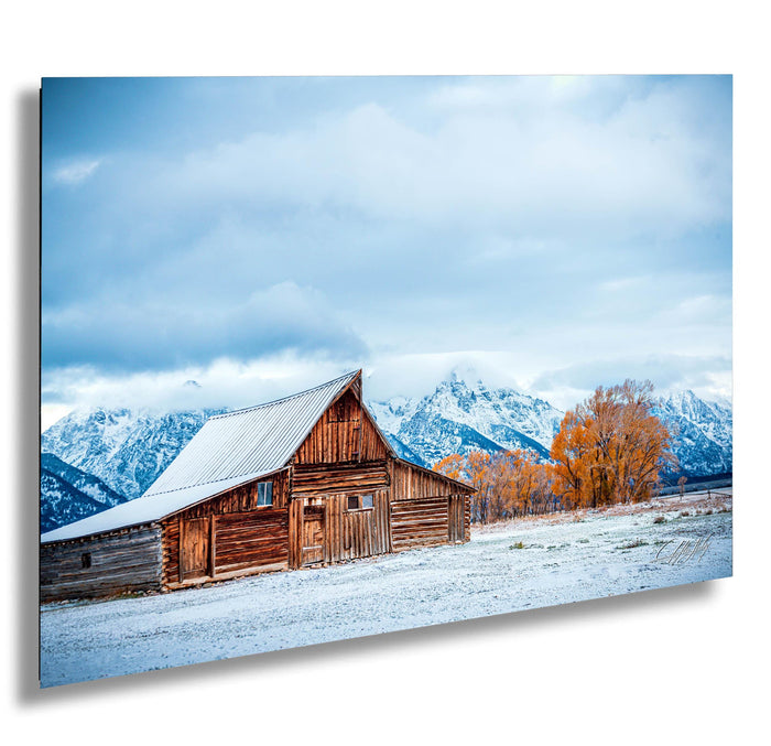 A rustic wooden barn stands in a snowy landscape, with snow-capped mountains in the background and a cloudy sky above.