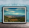 The image is a framed photograph of a scenic landscape, featuring a valley with a city in the distance, surrounded by mountains and trees.