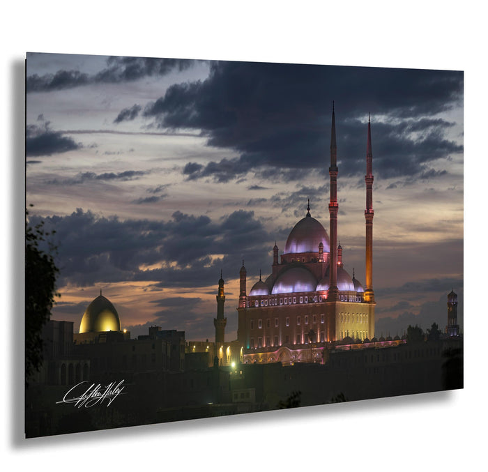 A large, illuminated mosque with multiple domes and minarets stands prominently against a dark, cloudy sky at dusk.