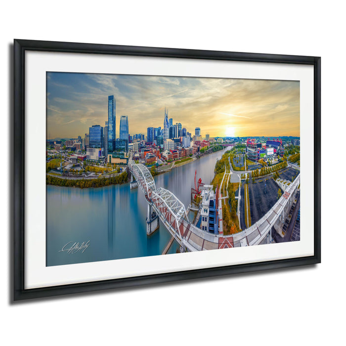 The image is a framed aerial view of a city skyline, featuring a bridge, a river, and a sunset in the background.