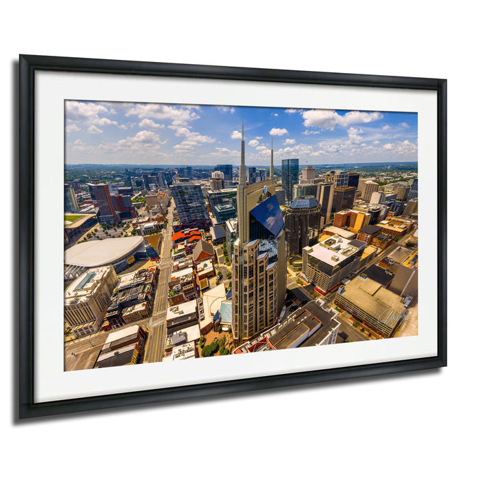 The image is a framed aerial view of a bustling city, showcasing a variety of tall buildings, skyscrapers, and other structures. The cityscape is vibrant and densely populated, with a mix of architectural styles and colors. The image is framed within a black border, and the overall composition is visually striking and dynamic.