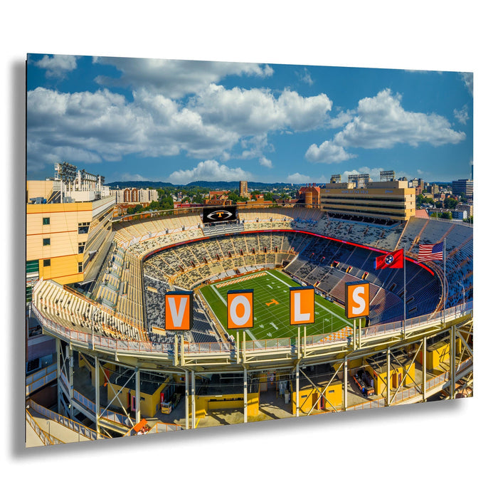 A large sports stadium with a football field, surrounded by buildings and a clear blue sky with clouds.