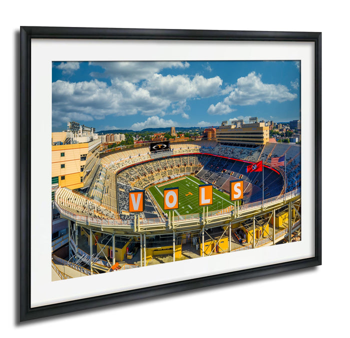 a large stadium with a football field, surrounded by buildings and a clear blue sky with clouds.