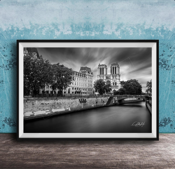 A black and white photograph of the Notre-Dame Cathedral in Paris, France, with a river and trees in the foreground.