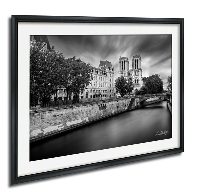 A black and white photograph of the Notre-Dame Cathedral in Paris, France, with a river running alongside the cathedral and surrounding buildings.