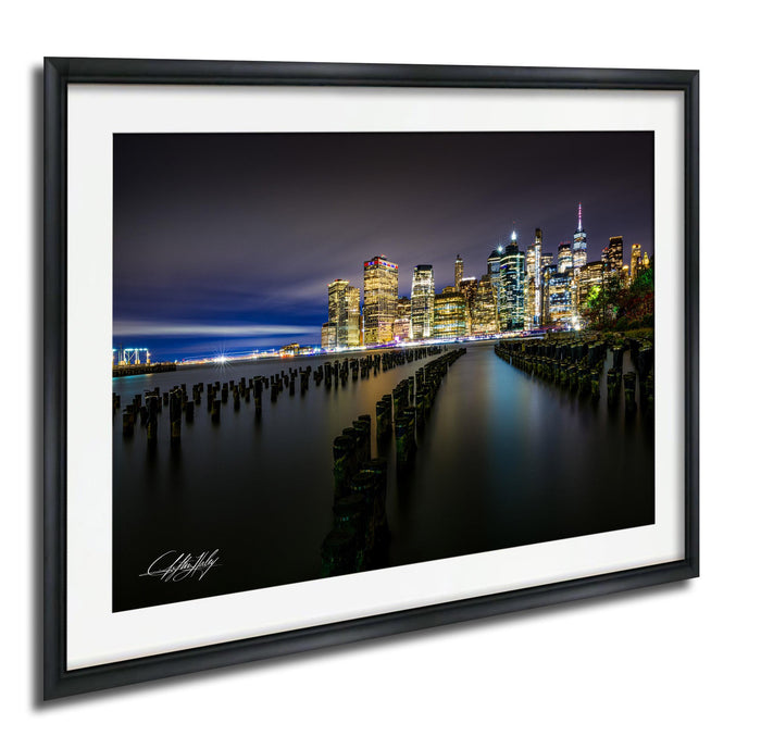 A nighttime cityscape with illuminated skyscrapers and a body of water in the foreground.