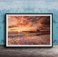 A framed photograph depicting a serene beach scene at sunset, with a wooden pier extending into the water and the sky filled with vibrant orange and pink hues.