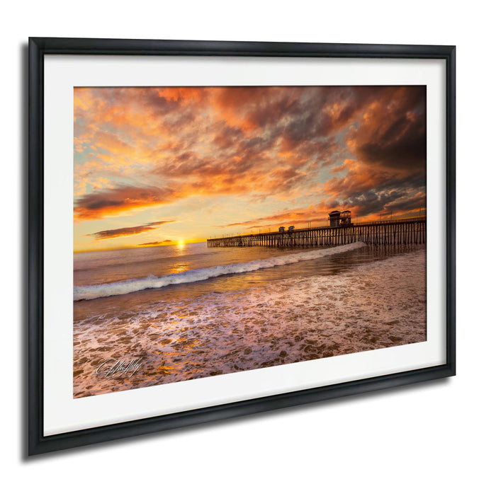 A framed photograph depicting a serene beach scene at sunset, with a wooden pier extending into the water and the sky filled with vibrant orange and pink hues.
