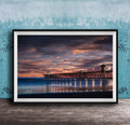 A framed photograph of a wooden pier extending into the ocean at sunset, with the sky filled with vibrant colors and clouds.