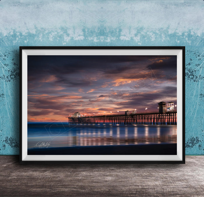 A framed photograph of a wooden pier extending into the ocean at sunset, with the sky filled with vibrant colors and clouds.