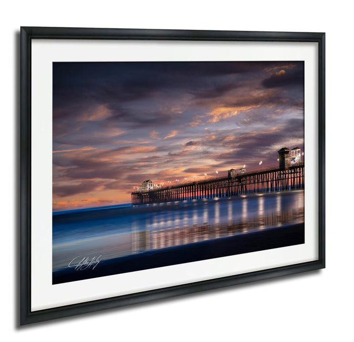 A framed photograph depicting a serene coastal scene at sunset, with a wooden pier extending into the calm waters and the sky filled with vibrant hues of orange and pink.