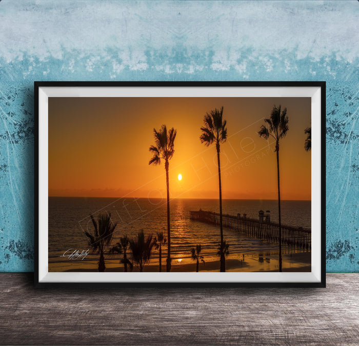 A framed photograph of a serene beach scene at sunset, with palm trees silhouetted against the warm orange sky.