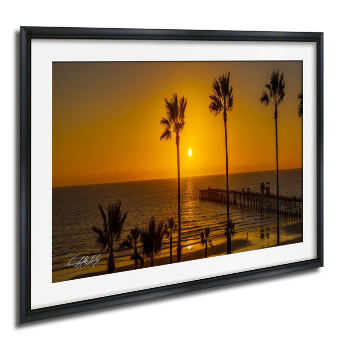 A framed photograph depicting a serene beach scene at sunset, with palm trees silhouetted against the warm, golden sky.