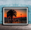 A framed photograph depicting a palm tree silhouetted against a vibrant sunset over the ocean.
