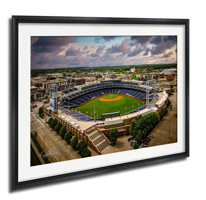 An aerial view of a baseball stadium with a lush green field, surrounded by buildings and a cloudy sky.