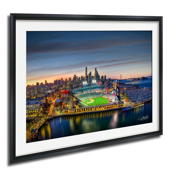 A framed aerial view of a city skyline at dusk, with a baseball field and stadium illuminated in the foreground.