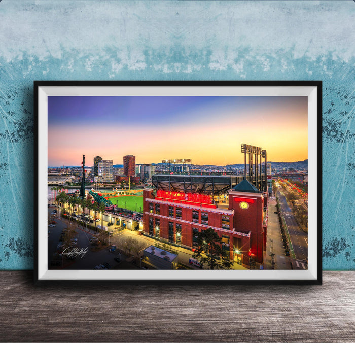A framed photograph of a city skyline at dusk, with a large stadium and surrounding buildings illuminated against a colorful sunset sky.