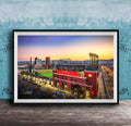 A framed photograph of a city skyline at dusk, with a large stadium and surrounding buildings illuminated against a colorful sunset sky.