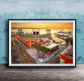 The image is a framed aerial view of a cityscape with a large stadium, buildings, and a body of water in the background.