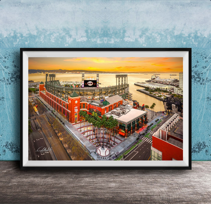 The image is a framed aerial view of a cityscape with a large stadium, buildings, and a body of water in the background.