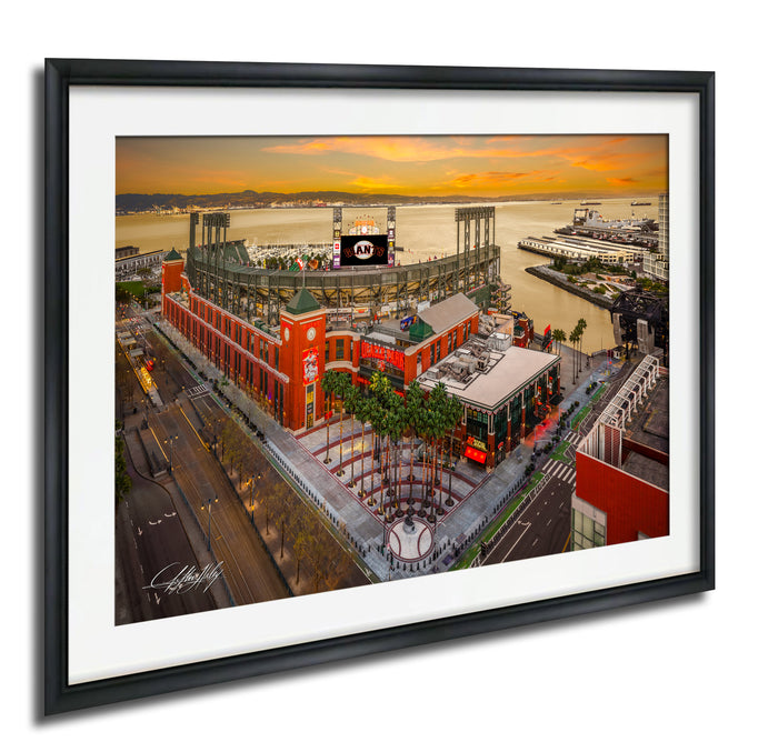 The image is a framed aerial view of a large stadium with a red and white exterior, surrounded by palm trees and other buildings. The stadium appears to be a sports venue, with a large screen visible in the background. The sky is filled with warm, orange and yellow hues, suggesting a sunset or sunrise. The image is framed with a black border, and the artist's signature is visible in the bottom left corner.