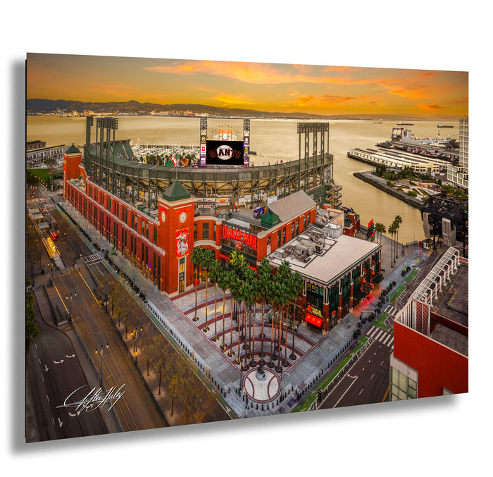 The image is an aerial view of a large stadium with a red and white exterior, surrounded by palm trees and other buildings. The stadium appears to be a sports venue, with a large scoreboard visible in the background. The sky is filled with warm, orange and yellow hues, suggesting a sunset or sunrise. The image is a collage of multiple images, and the artist's signature is visible in the bottom left corner.