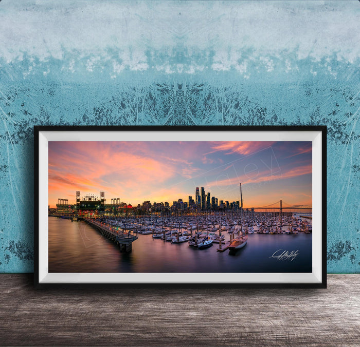 A framed photograph depicting a city skyline at sunset, with boats docked in a harbor and a large stadium in the background.