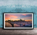 A framed photograph depicting a city skyline at sunset, with boats docked in a harbor and a large stadium in the background.