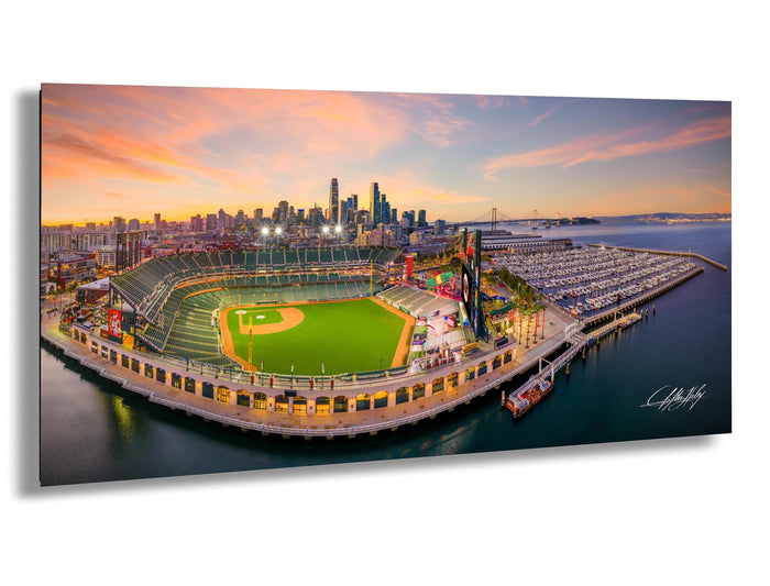 An aerial view of a baseball stadium with a city skyline in the background, set against a colorful sunset sky.