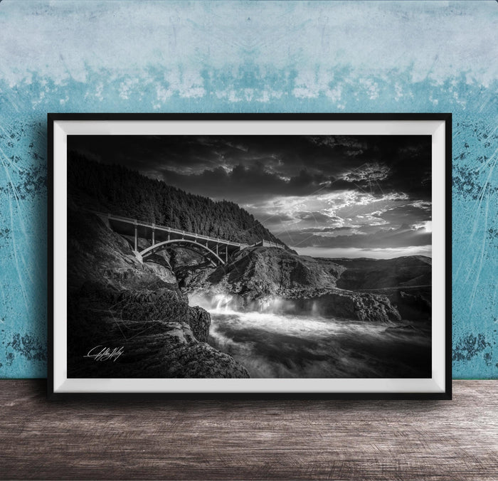 A black and white photograph of a bridge spanning over a rushing river, with dramatic clouds in the sky above.