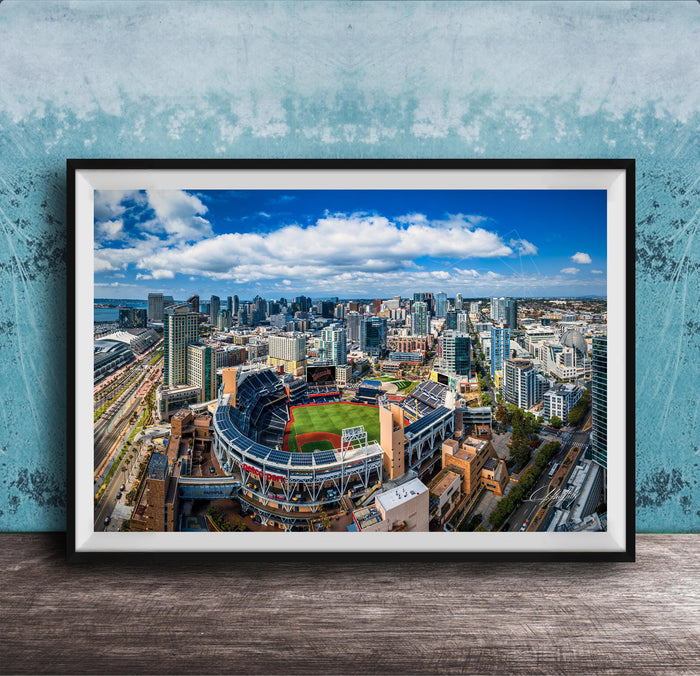 An aerial view of a large city with a baseball stadium in the center, surrounded by tall buildings and a blue sky with clouds.