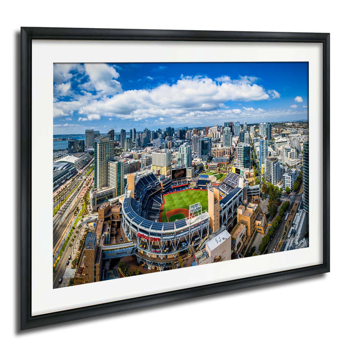 An aerial view of a large city with a baseball stadium in the center, surrounded by tall buildings and a clear blue sky with white clouds.