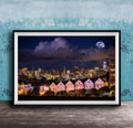 A framed photograph of a city skyline at night, with a full moon in the sky and colorful houses lining the streets.