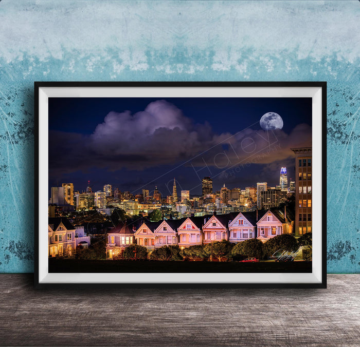 A framed photograph of a city skyline at night, with a full moon in the sky and colorful houses lining the streets.