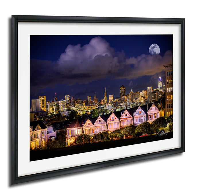 a cityscape at night, with a full moon illuminating the skyline and a row of houses with their lights on in the foreground.