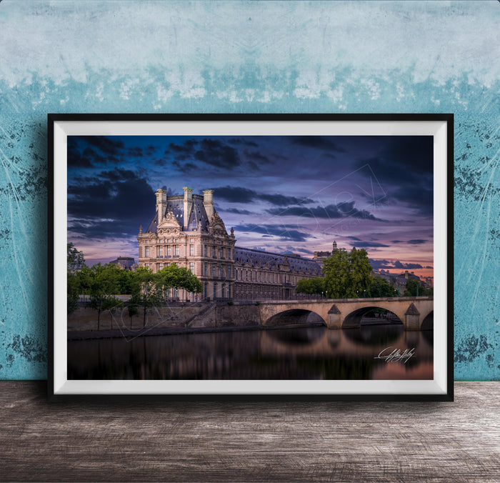 a large, ornate building with a tower and multiple spires, situated on the banks of a river. The sky is filled with clouds, creating a dramatic backdrop for the scene. The building is surrounded by trees, and a bridge can be seen in the background. The image is framed and displayed on a wooden surface, with a blue wall in the background.