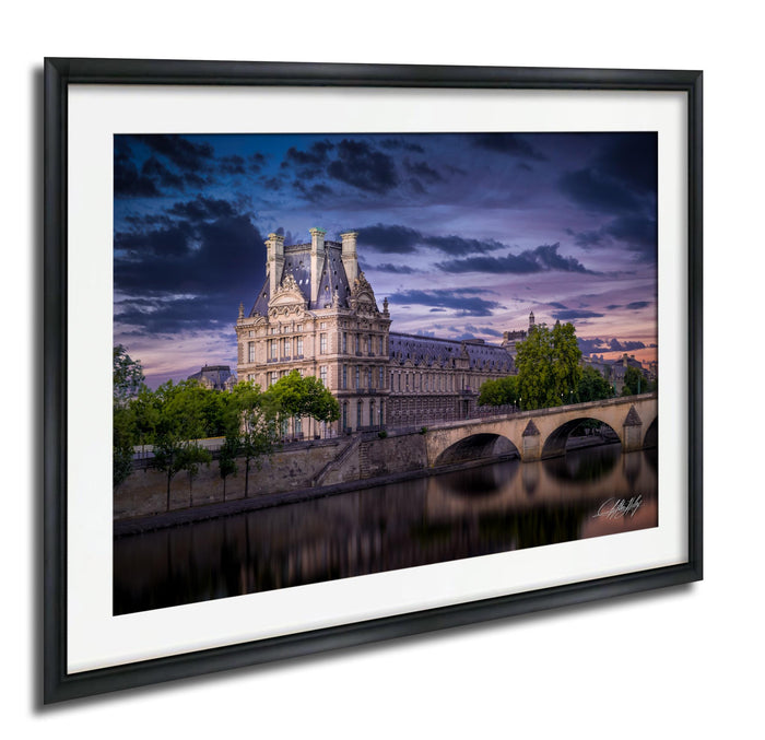 a large, ornate building with a tower and multiple spires, situated along a river with a bridge in the background. The sky is filled with clouds, creating a dramatic atmosphere.