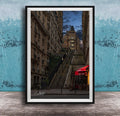 a city street with a tall, ornate building on a steep, winding staircase. The building has a distinctive dome and is surrounded by other buildings. The street is lined with trees and has outdoor seating areas, suggesting a cafe or restaurant. The sky is dark and cloudy, creating a moody atmosphere. The image is framed and displayed on a wooden surface, with a blue wall in the background.