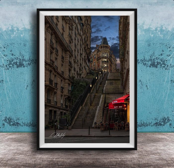 a city street with a tall, ornate building on a steep, winding staircase. The building has a distinctive dome and is surrounded by other buildings. The street is lined with trees and has outdoor seating areas, suggesting a cafe or restaurant. The sky is dark and cloudy, creating a moody atmosphere. The image is framed and displayed on a wooden surface, with a blue wall in the background.