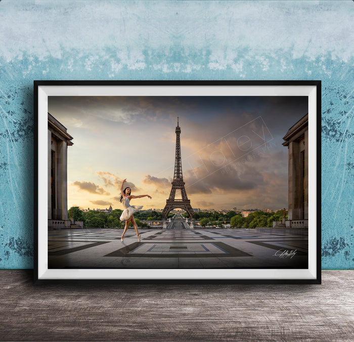 A framed photograph of a woman in a white dress dancing in front of the Eiffel Tower in Paris, France, with a blue sky and clouds in the background.
