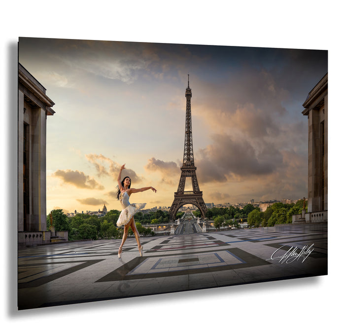 A woman in a white tutu is dancing in front of the Eiffel Tower in Paris, France, with a dramatic sky in the background.