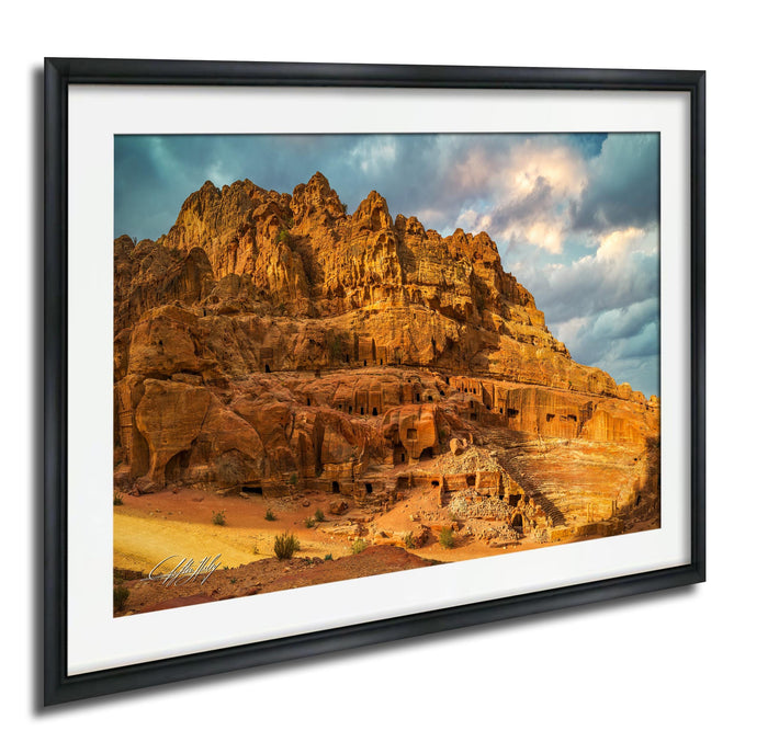 a large, rocky cliff with a rugged, reddish-brown surface. The cliff is covered in various rock formations and features, creating a dramatic and visually striking landscape. The sky above the cliff is filled with clouds, adding to the dramatic effect of the scene. The image is framed and appears to be a photograph of the rock cliff, showcasing its unique and captivating features.