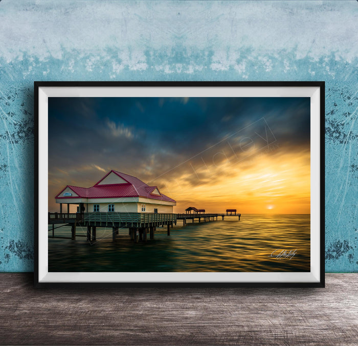 A framed photograph depicting a serene pier scene at sunset, with a red-roofed building and a wooden pier extending into the calm waters.